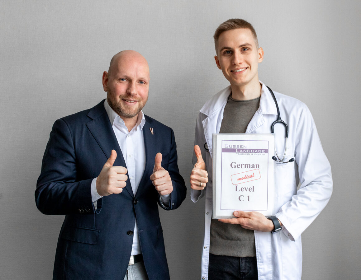 CEO Sascha Gussen and a medical professional who is a graduate of our scholarship program. They are standing next to each other and smiling, with thumbs up. The medical professional is wearing a white lab coat, a stethoscope around his neck, and holding a C1 German language certificate in his hands."